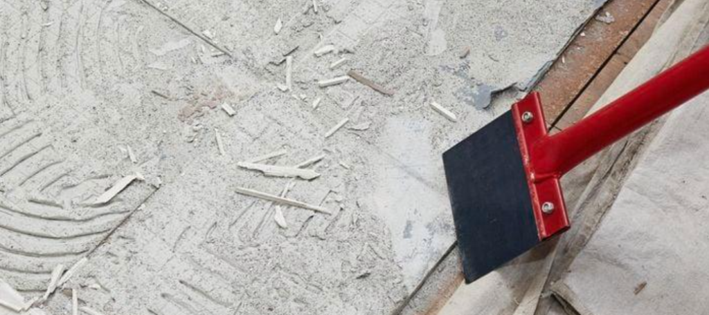 Using a floor scraper to lift a loose edge of old tile adhesive and check its texture and flexibility.